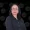 Portrait of a woman with dark hair and glasses wearing a black button down shirt posed against a black backdrop with circular portfolio watermarks