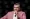Man in a pink blazer speaks into a microphone on stage during an event