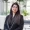 Professional portrait of a woman with long dark hair in a black blazer standing in a bright office lobby with plants in the background