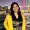 Smiling woman with shoulder length dark hair wearing a yellow jacket over a black and gold dress standing by the water with a lit bridge and city skyline behind