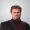 Professional headshot of a man with brown hair and goatee wearing a black blazer and burgundy turtleneck against a gray backdrop