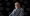 Professional portrait of a man wearing glasses a gray plaid suit and scarf seated against a black background with watermark patterns reading The Portfolio